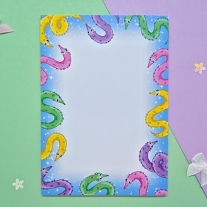 May include: A rectangular sheet of stationery featuring a border of cartoon worms in various colours, including yellow, pink, purple, and green, against a light blue backdrop. The stationery is set on a green and purple surface.