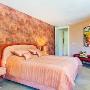 May include: A bedroom with a large bed with a pink comforter and a headboard with a pink upholstered headboard. The bed has several pillows in shades of pink and peach. There is a nightstand with a lamp next to the bed. The wall behind the bed is covered in a brown and gold wallpaper with a leaf pattern. There is a door to the right of the bed and a small sofa with pillows in the background.