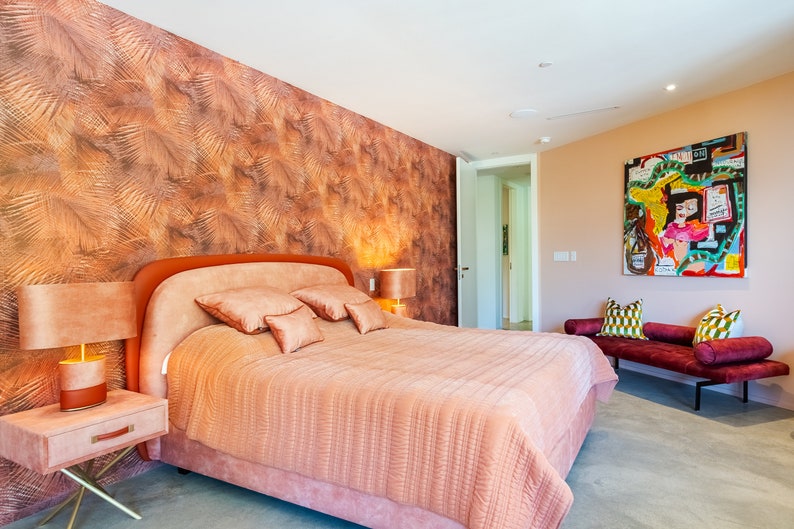 May include: A bedroom with a large bed with a pink comforter and a headboard with a pink upholstered headboard. The bed has several pillows in shades of pink and peach. There is a nightstand with a lamp next to the bed. The wall behind the bed is covered in a brown and gold wallpaper with a leaf pattern. There is a door to the right of the bed and a small sofa with pillows in the background.