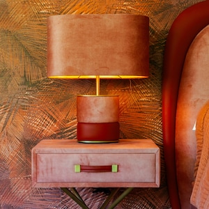 May include: A bedside table with a drawer and a lamp. The table and lamp are both a dusty rose color. The lamp has a round, light brown shade. The table has a gold metal base.