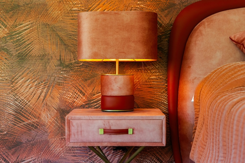 May include: A bedside table with a drawer and a lamp. The table and lamp are both a dusty rose color. The lamp has a round, light brown shade. The table has a gold metal base.