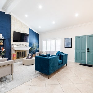 May include: A living room interior with a blue and white color scheme. The room features a fireplace with a white mantel, a large flat-screen TV, a blue velvet sofa, and two gray armchairs. The room has a white ceiling and white walls with a blue accent wall. The floor is tiled in a light beige color.