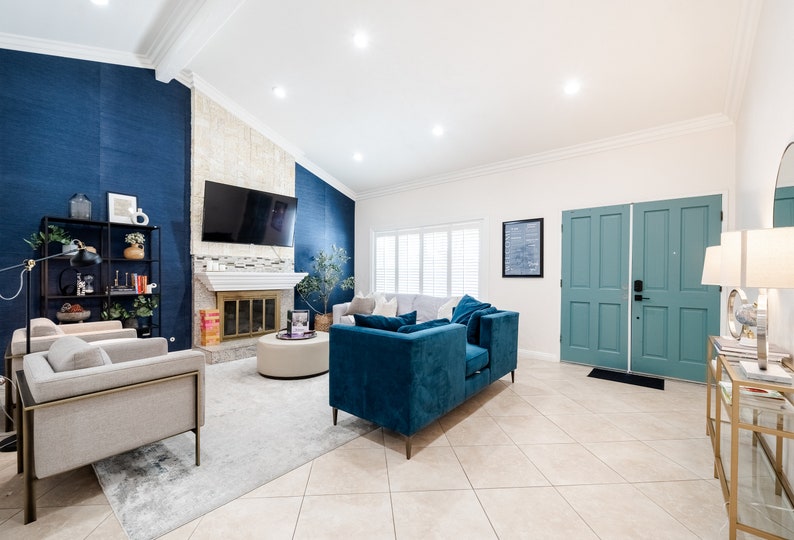 May include: A living room interior with a blue and white color scheme. The room features a fireplace with a white mantel, a large flat-screen TV, a blue velvet sofa, and two gray armchairs. The room has a white ceiling and white walls with a blue accent wall. The floor is tiled in a light beige color.