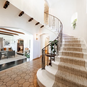 May include: A white staircase with a wrought iron railing curves up to a landing. The stairs are carpeted in a light brown color. The floor is tiled in a hexagonal pattern.