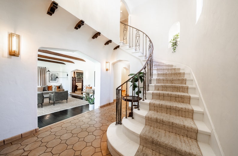 May include: A white staircase with a wrought iron railing curves up to a landing. The stairs are carpeted in a light brown color. The floor is tiled in a hexagonal pattern.