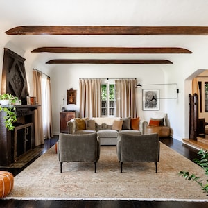 May include: A living room with a white fireplace, a large rug, and two gray armchairs. The room has exposed wooden beams and a large window with beige curtains.