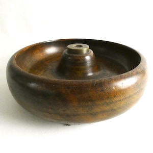 May include: A circular wooden bowl with a central, raised brass fitting. The bowl is a dark brown colour with visible wood grain. The bowl's design is simple and functional, with a wide, shallow basin.