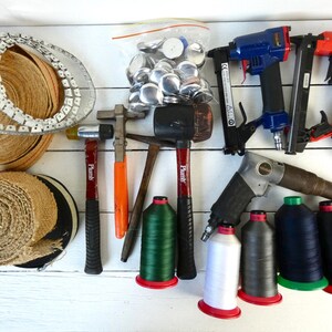 May include: A collection of upholstery tools and supplies including two rolls of jute webbing, a metal tack strip, a bag of metal buttons, two pneumatic nail guns, a pneumatic stapler, three hammers, and six spools of thread in white, grey, green, blue, black, and tan.