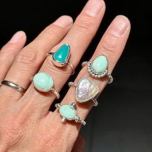 May include: A collection of five silver rings featuring various gemstones. One ring has a smooth silver band, while others showcase teardrop, oval, and heart-shaped stones in shades of turquoise, mint green, and iridescent white set in silver.