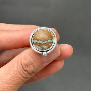 May include: A circular ring featuring a polished brown stone with a horizontal turquoise vein running through the center. The stone is set within a smooth silver bezel, framed by a thin silver wire border, and accented with a small silver bead.