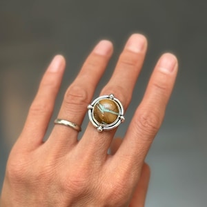 May include: A silver ring featuring a round, polished brown stone with distinct turquoise-colored veining. The stone is set in a smooth silver bezel, accented by four small silver spheres positioned at the north, south, east, and west points.