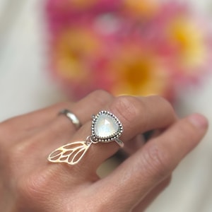 Puede incluir: Anillo de plata con una piedra preciosa triangular iridiscente y un dije de ala calada. La piedra preciosa está rodeada por un borde de cuentas. El anillo se lleva en un dedo, con un fondo floral borroso.