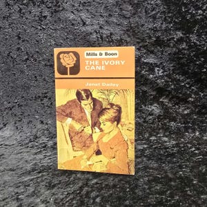 May include: A vintage paperback book titled "The Ivory Cane" by Janet Dailey. The cover features a couple in a romantic scene, with the title and author's name in a bold font. The book is from the Mills & Boon collection.