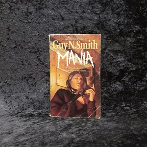 May include: A paperback book cover with a dark red background and a woman with dark hair and a red jacket holding a knife. The title of the book is "MANIA" by Guy N. Smith. The text on the cover reads "A living nightmare on the edge of madness."