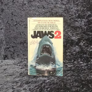 May include: Vintage paperback book titled "JAWS 2" featuring a menacing shark illustration on the cover. The cover has text that reads "A Completely New Novel by Hank Searls". The book is set against a dark background.