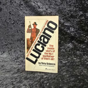 May include: A vintage paperback book titled "Luciano" with a cover illustration of a man in a suit holding a gun. The book's title is in large black letters, with the subtitle "The inside story of the No.1 Godfather of them all!"