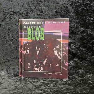 May include: A hardcover book titled "Famous Movie Monsters Meet The Blob" by Suzanne J. Murdico. The cover features the word "BLOB" in green, outlined in black, against a dark, textured background with white accents and a pink border.