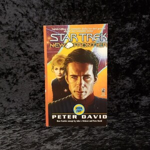 May include: A Star Trek: New Frontier book cover featuring a yellow, orange, and blue background with a space scene. The cover features a portrait of a man and a woman in Starfleet uniforms. The title "Star Trek: New Frontier" is in large, bold letters. The text "Book Two" is in a circle with a blue background. The author's name, "Peter David", is in large, bold letters. The text "New Frontier concept by John J. Ordover and Peter David" is in smaller letters below the author's name.