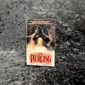 May include: A paperback book titled "The Piercing" by John Coyne. The cover features a woman in a bathtub with arms raised, and the title in red. The book is a Berkley Book.