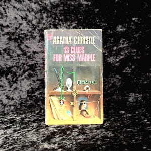 May include: A paperback book cover with a brown background and a yellow border. The title is "13 Clues for Miss Marple" by Agatha Christie. The cover art features a shelf with a key, an egg, a needle, and a bloody eye.