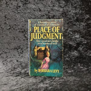 May include: A paperback book cover with a dark blue background and a spooky house with a woman in a pink dress standing in front of it. The title of the book is "Place of Judgment" and the author is Barbara Levy.