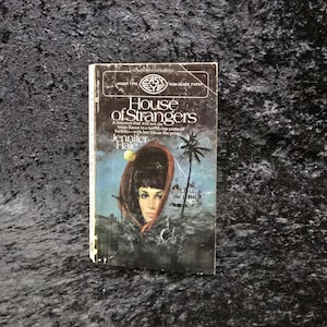 May include: A vintage paperback book titled "House of Strangers" by Jennifer Hale. The cover features a woman's portrait within a dark frame, a palm tree, and a white building. The book has a worn appearance.