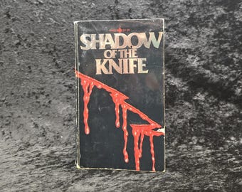 May include: A vintage paperback book titled "Shadow of the Knife." The cover is black with the title in silver lettering. A red, blood-like design drips down the front. The book has a price tag of $1.95.