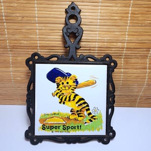 May include: Black cast iron trivet with a square ceramic tile featuring a cartoon tiger playing baseball. The tiger is yellow and black, wearing a blue baseball cap, and holding a bat. The tile reads "Super Sport!" and "© 1976 Suzy's Zoo."