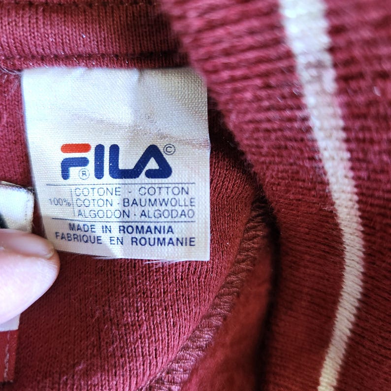 May include: Close-up of a burgundy garment with a white stripe. A white label displays the FILA logo in blue and red, along with text in multiple languages: Italian, English, French, German, Portuguese, and Spanish. The label reads "Made in Romania."