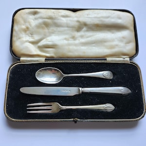 May include: A silver cutlery set comprising a spoon, knife, and fork, displayed in a black case with a cream satin lining. The antique silverware features decorative detailing. The case is rectangular.
