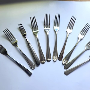 Può includere: Una collezione di forchette d'argento antiche disposte su una superficie bianca. Le forchette variano nel design, con diversi stili di manico e motivi decorativi. I rebbi sono distanziati uniformemente.
