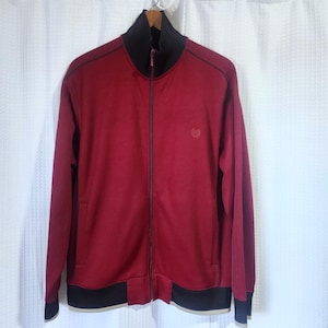 May include: A burgundy zip-up track jacket with a black collar, cuffs, and hem. The jacket has a small emblem on the chest and black stitching details. The jacket is hanging on a wooden hanger.