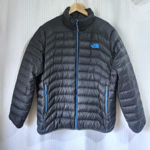 Peut inclure: Une doudoune matelassée noire dotée d'un col montant, d'une fermeture éclair centrale sur toute la longueur et de deux poches latérales zippées. Le logo The North Face bleu vif est visible sur la poitrine gauche avec des tirettes bleues.