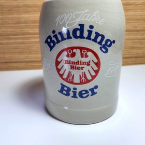 May include: A light grey ceramic beer stein with the words "Binding Bier" in blue and red. The stein features a red and white emblem with an eagle design and the text "100 Jahre" in white.