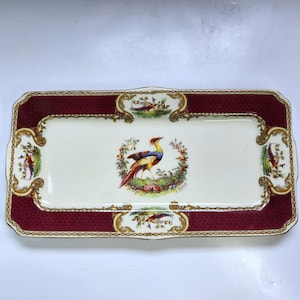 May include: Rectangular serving tray with a cream-colored center and a burgundy border. The tray features a colorful bird illustration surrounded by floral accents. Gold trim and decorative corner designs complete the elegant look.