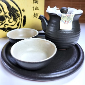 May include: A black ceramic tea set comprising a teapot, two bowls, and a tray. The teapot features a rounded body, a spout, and a wooden knob on the lid. The bowls have a light interior and dark rims. The set rests on a black tray.