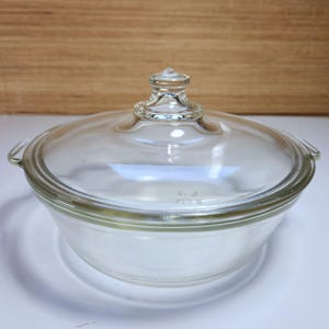 May include: A clear glass Pyrex bowl with a matching lid. The bowl has a rounded shape with small handles on either side. The lid has a knob handle. The bowl and lid are transparent, allowing visibility of the contents.