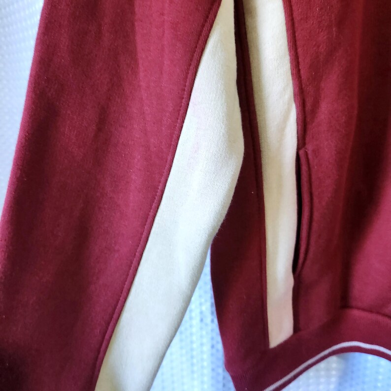 May include: Close-up of a maroon sweatshirt with a cream-colored stripe down the sleeve and side. The sweatshirt has a pocket and a white trim along the bottom.