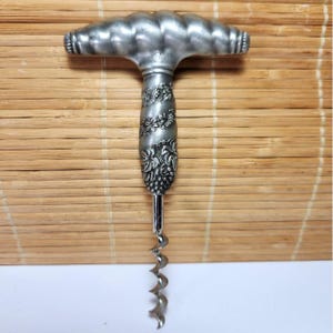 May include: A silver corkscrew with a decorative handle. The handle features a ridged top and a detailed floral and grape design on the body. The corkscrew's spiral is visible below the handle.