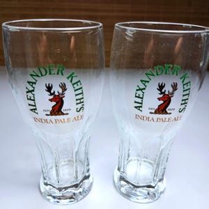以下が含まれることがあります： 「Alexander Keith's」のロゴ、鹿のエンブレム、そして「India Pale Ale」が描かれた、透明なビールグラス2つ。グラスはチューリップ型で、ビール用です。