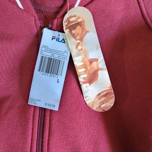 May include: A maroon FILA fleece zip-up hoodie with white stripes on the collar. The tag features a picture of a man playing a sport and the FILA logo. The tag also includes the size, style number, and barcode.
