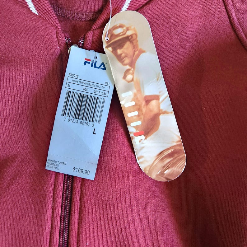 May include: A maroon FILA fleece zip-up hoodie with white stripes on the collar. The tag features a picture of a man playing a sport and the FILA logo. The tag also includes the size, style number, and barcode.