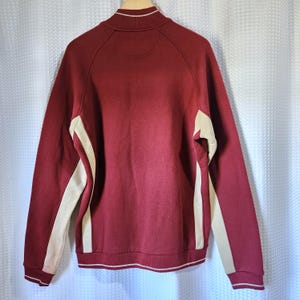 May include: Maroon zip-up sweatshirt with cream-colored side panels and a white stripe detail at the hem and collar. The back of the sweatshirt is visible, showcasing a minimalist design. The sweatshirt has a classic, sporty aesthetic.
