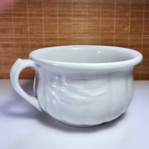 May include: A white ceramic shaving mug with a handle and a decorative leaf pattern. The mug has a wide opening and a curved rim. The mug is sitting on a white surface.