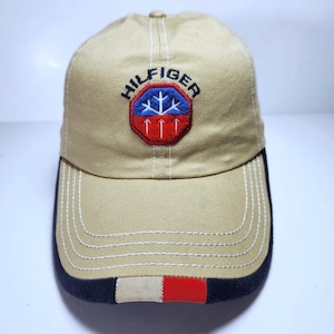 May include: A tan baseball cap featuring a central embroidered patch with the text HILFIGER above a blue snowflake and three white arrows on a red background. The brim has white stitching, a dark navy edge, and a small rectangular red and white accent.