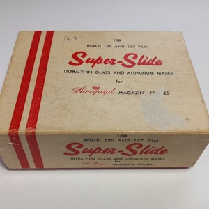 May include: A vintage box for Super-Slide film, featuring a cream-colored background with red stripes and text. The box reads "Super-Slide" in a bold, red font, with additional text indicating the film type and its use with magazine frames.