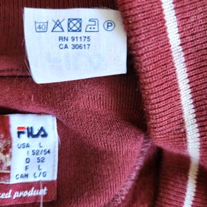 May include: Close-up of a maroon-colored garment with white stripes. The garment has a tag with washing instructions and the brand "FILA". The tag also indicates the size as L (52/54).