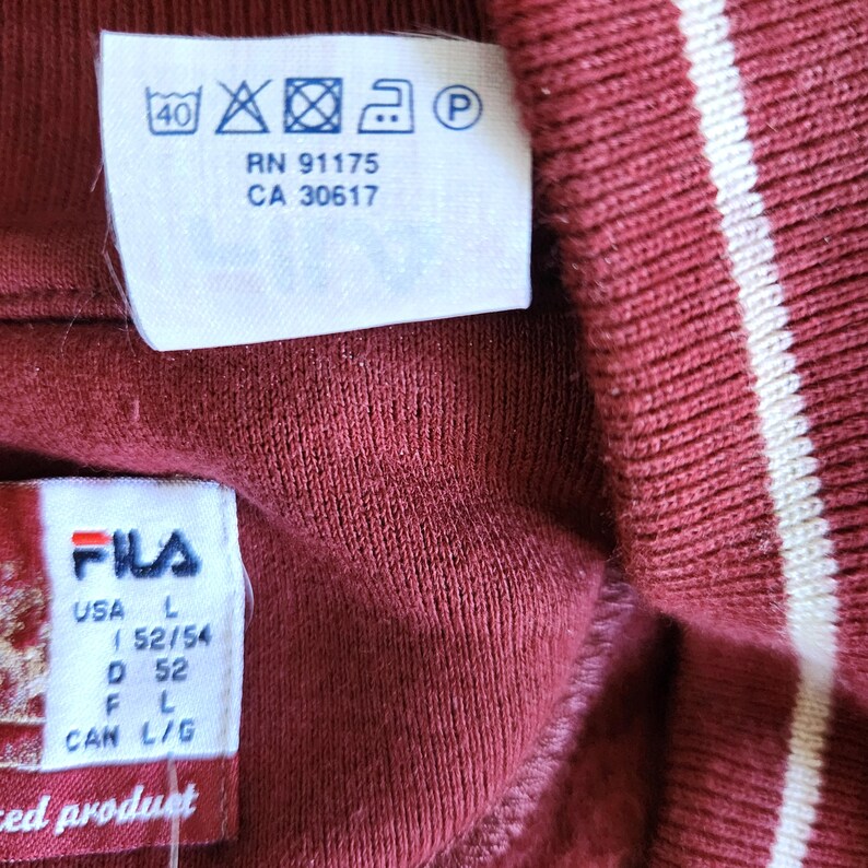 May include: Close-up of a maroon-colored garment with white stripes. The garment has a tag with washing instructions and the brand "FILA". The tag also indicates the size as L (52/54).