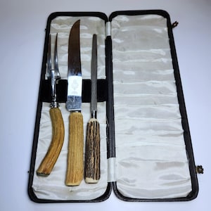 Può includere: Un set da intaglio composto da una forchetta a due rebbi, un coltello da intaglio e un acciaino, tutti dotati di manici in corno testurizzato. Il set è contenuto in una custodia rettangolare incernierata, foderata in tessuto di raso bianco.