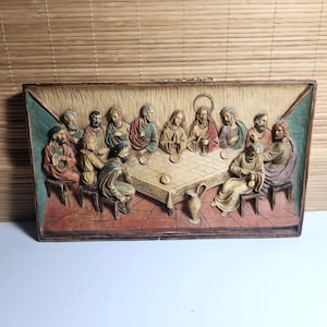 May include: A rectangular wall plaque featuring a relief carving of The Last Supper. The scene depicts thirteen figures seated around a rectangular table with a checkered tablecloth, rendered in muted tones of brown, red, green, and cream.